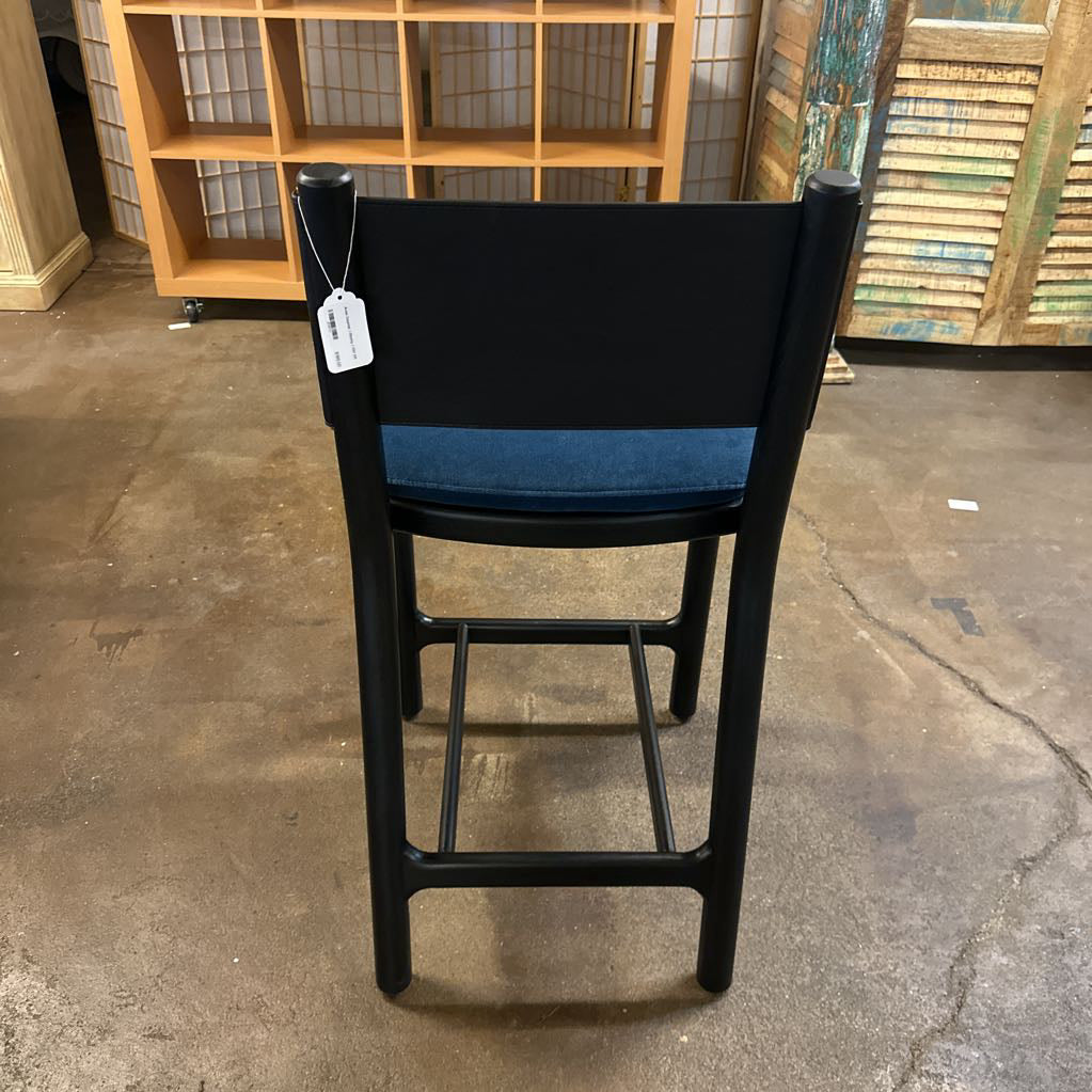 Black bar stool with blue seat cushion on a concrete floor, with a wooden shelf in the background.