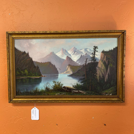Framed painting of a mountain landscape with a lake on an orange wall.