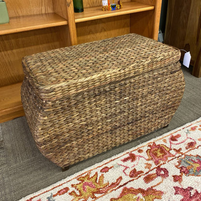 Woven storage ottoman on a carpeted floor with a wooden shelf in the background.