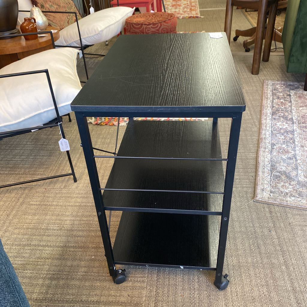 Black metal rolling cart with three shelves in a room setting.