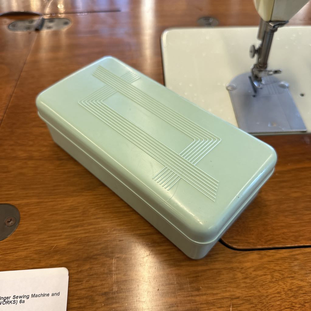 Light green sewing machine accessory box on a wooden surface with a sewing machine in the background.