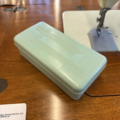 Light green sewing machine accessory box on a wooden surface with a sewing machine in the background.