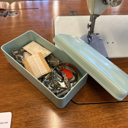 Container with sewing machine needles on a wooden table next to a sewing machine.