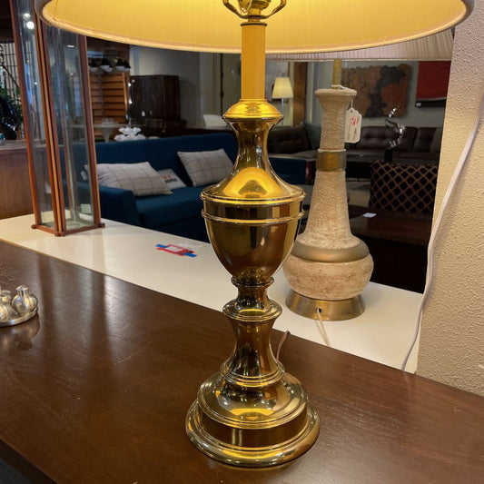 Gold table lamp with a yellow shade on a wooden surface, with a blurred indoor setting in the background.