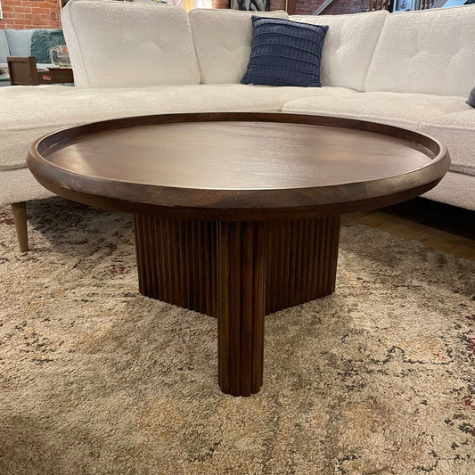 A round dark wood coffee table with a raised tray-style edge and a fluted, pedestal base, resting on a neutral-toned patterned rug
