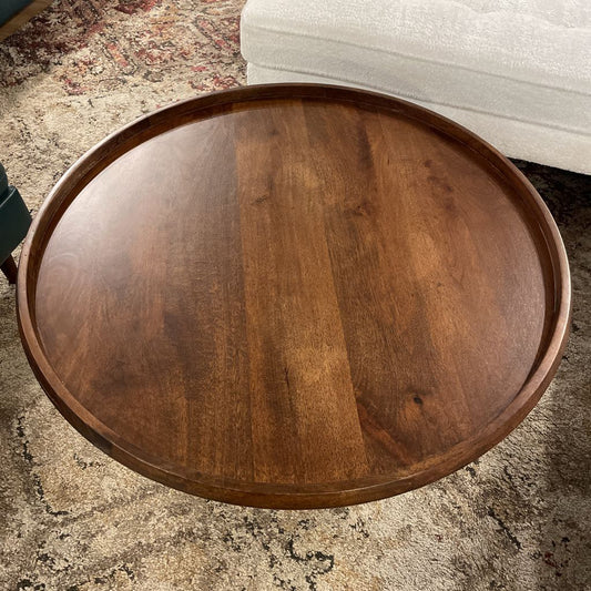 A round dark wood coffee table with a raised tray-style edge and a fluted, pedestal base, resting on a neutral-toned patterned rug