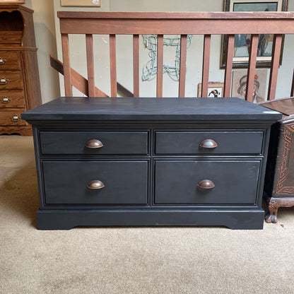 Black dresser with four drawers and brass handles in a room with a staircase and furniture.
