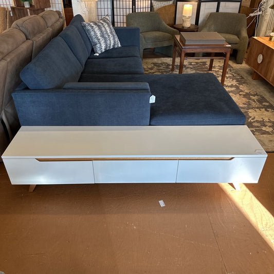 A long, white low-profile media console with three drawers, featuring integrated wooden pull-handles and tapered legs