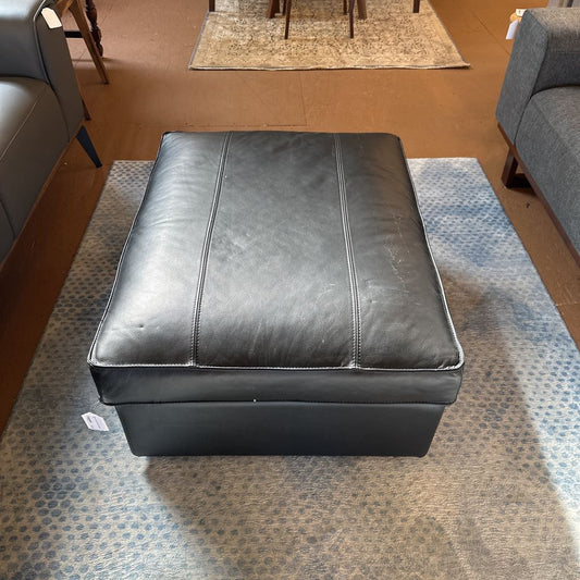 Black leather ottoman on a patterned rug in a room with furniture.