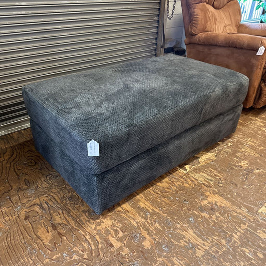 Gray ottoman with a price tag on a wooden floor next to a brown chair.