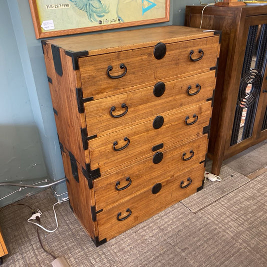 A rustic wooden Tansu-style chest with five drawers, featuring decorative black metal corner hardware, circular lock plates, and iron bail handles.