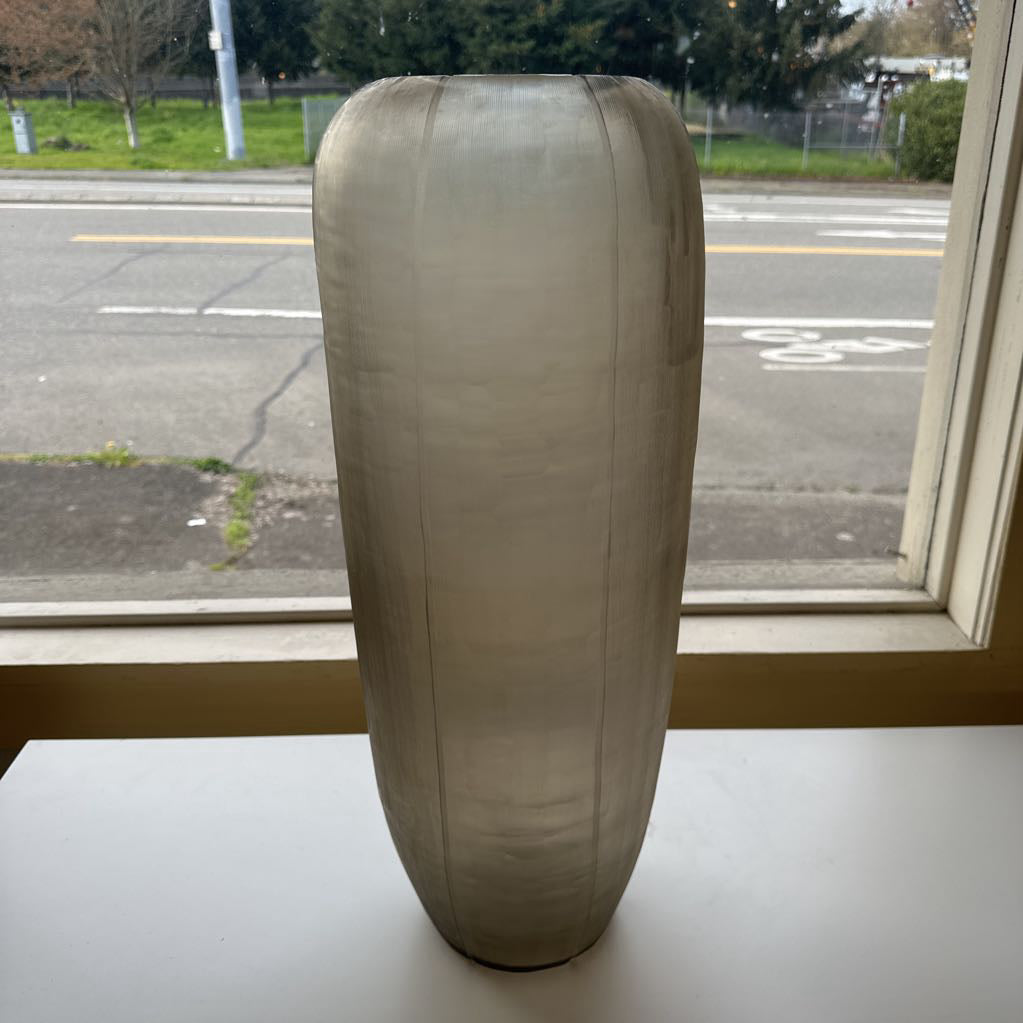 Large beige vase on a windowsill with a view of a road and trees outside.