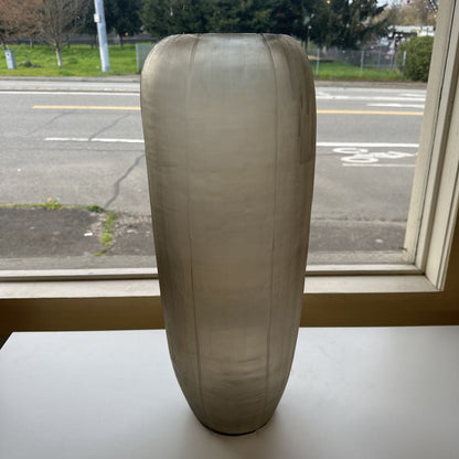 Large beige vase on a windowsill with a view of a road and trees outside.