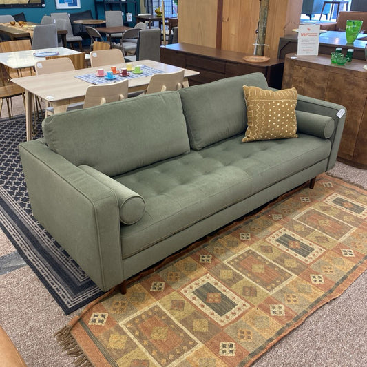 Green sofa with a patterned pillow on a decorative rug in a furniture store.