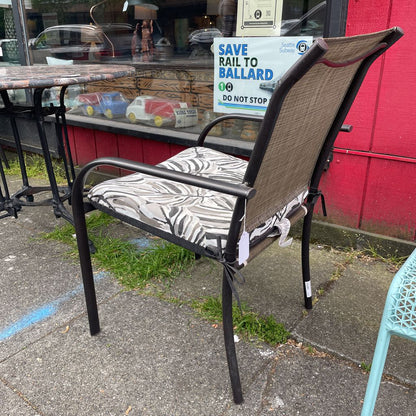 Angled side view of an outdoor patio chair with a black frame, grey mesh back, and tropical leaf print cushion