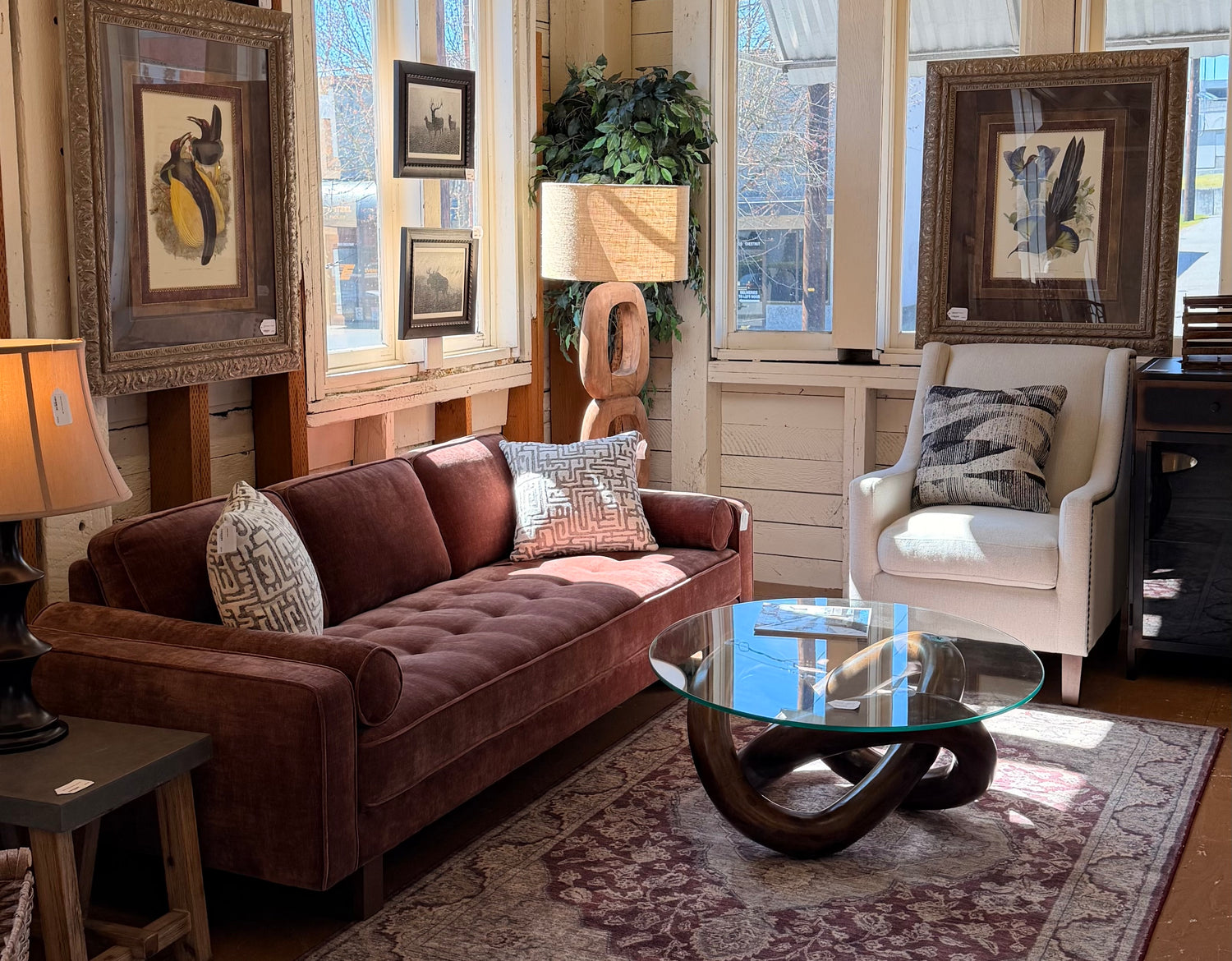 A staged living room set featuring a chocolate brown button-tufted velvet sofa, a cream wingback armchair, and a round glass coffee table with a sculptural wooden base, all arranged on a traditional patterned rug