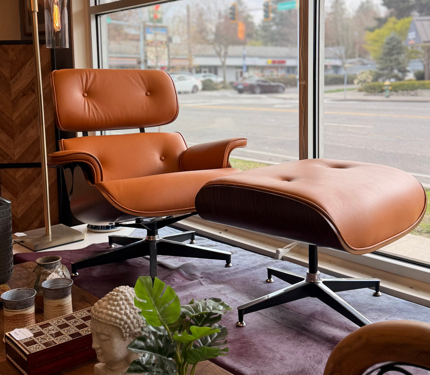 A mid-century modern lounge chair and matching ottoman in tan leather with dark wood shells, positioned in front of a large window overlooking a street.